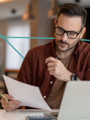 man with glasses and dark hair sitting near computer reading a paper document